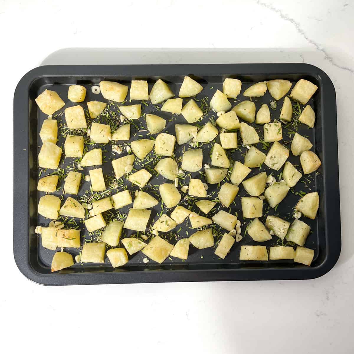 Partially cooked parmentier potatoes on a baking tray, sprinkled with garlic and rosemary.