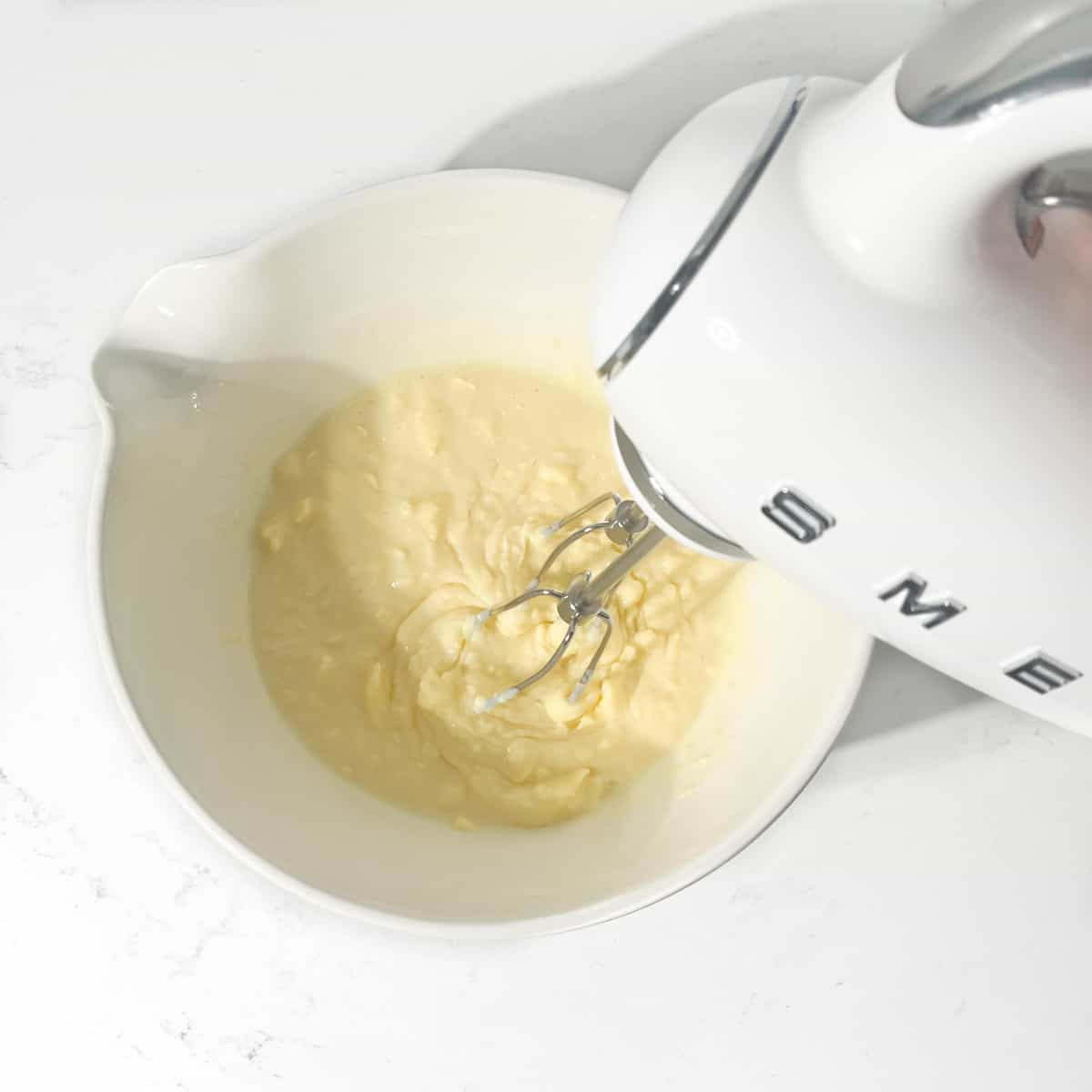 Mixing butter and condensed milk with an electric hand mixer in a large white bowl.