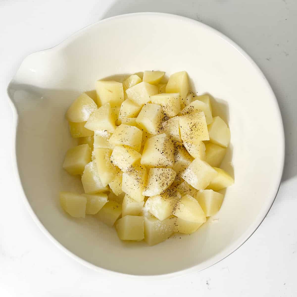 Potato cubes in a large white bowl with olive oil, salt and pepper.