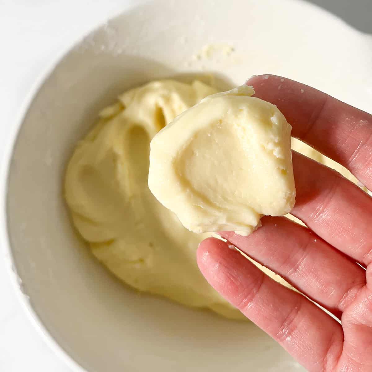 A piece of pinched off cookie dough held above a large white bowl.