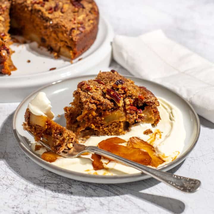 A slice of spiced apple cake on a light grey plate with some cream and caramelised apples.