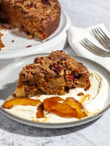 A slice of spiced apple cake on a light grey plate with some cream and caramelised apples.