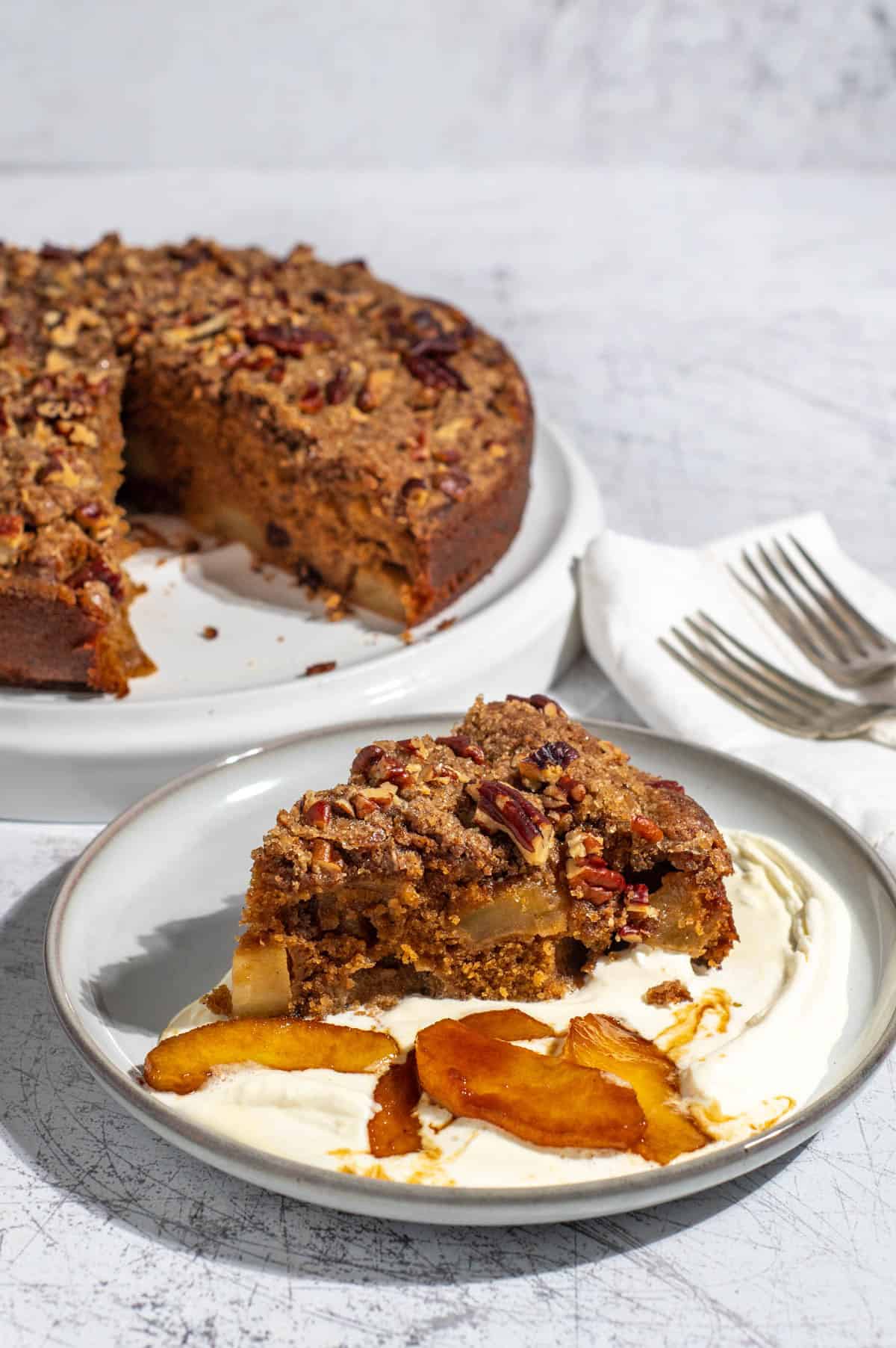 A slice of spiced apple cake on a light grey plate with some cream and caramelised apples.