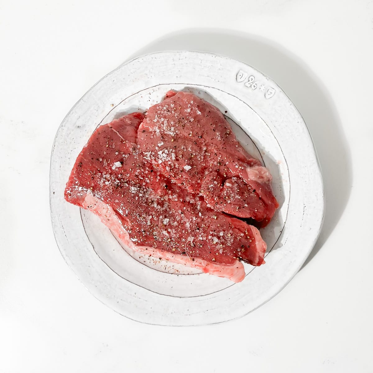 Raw steak on a plate that's been seasoned with salt and pepper. 