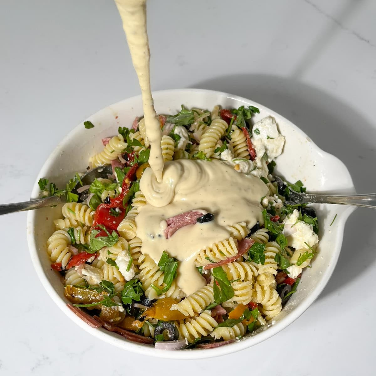 Adding the salad dressing to the cold pastas salad in a large white bowl.