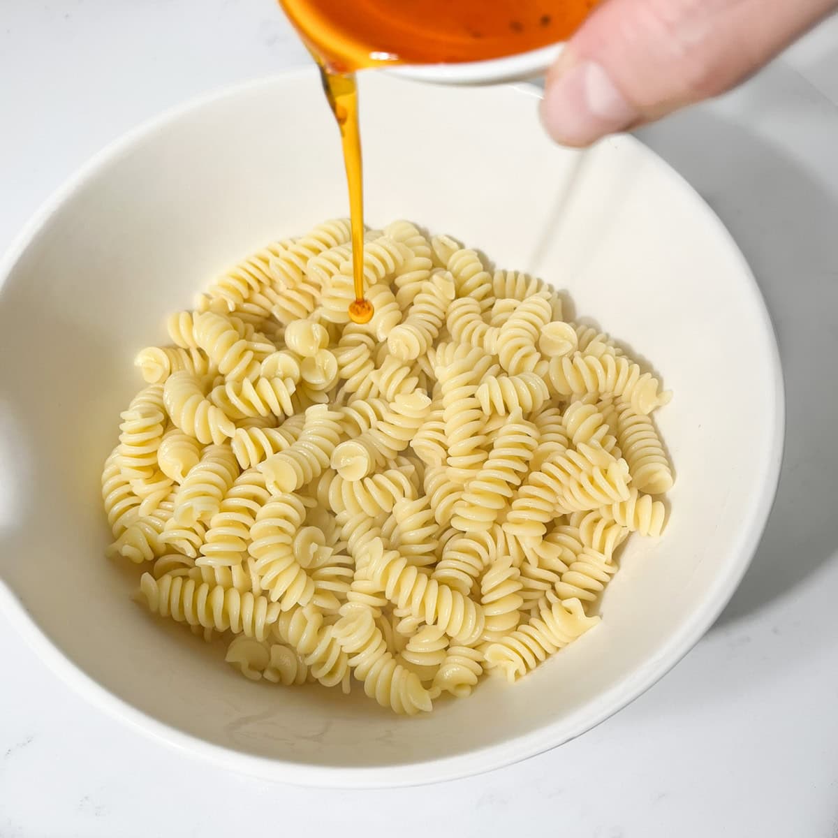 Adding some of the reserved red pepper oil to the cooked pasta in a large white bowl.