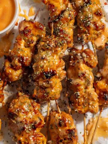 Bang bang chicken skewers on a white background with a small pot of bang bang sauce and lime wedges.
