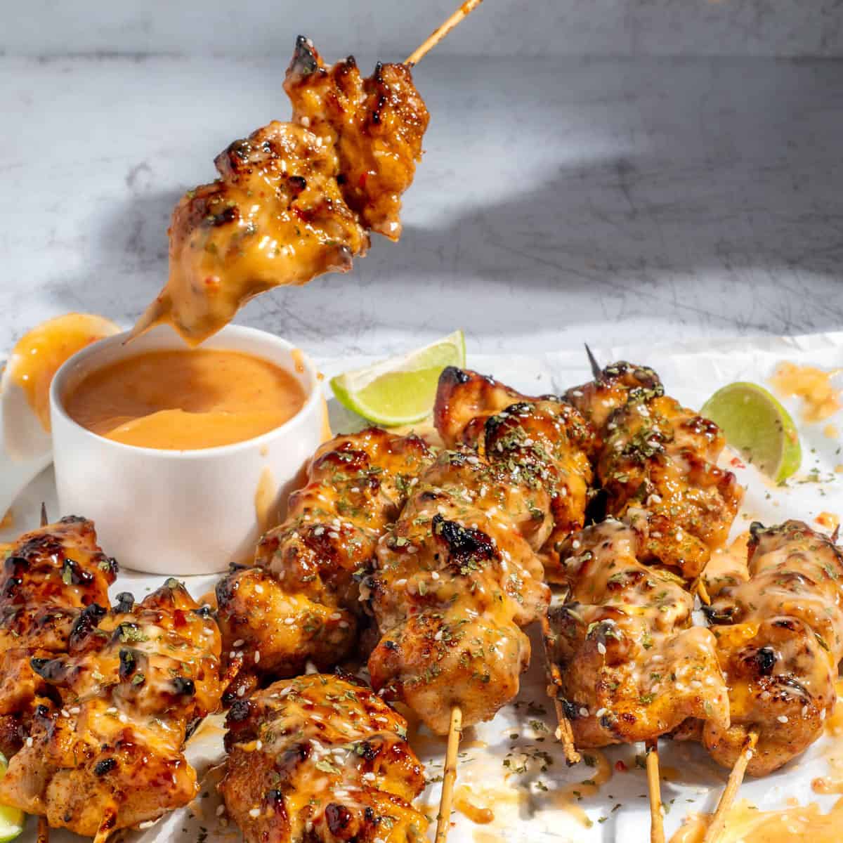 Bang bang chicken skewers on a white background with a small pot of bang bang sauce and lime wedges.