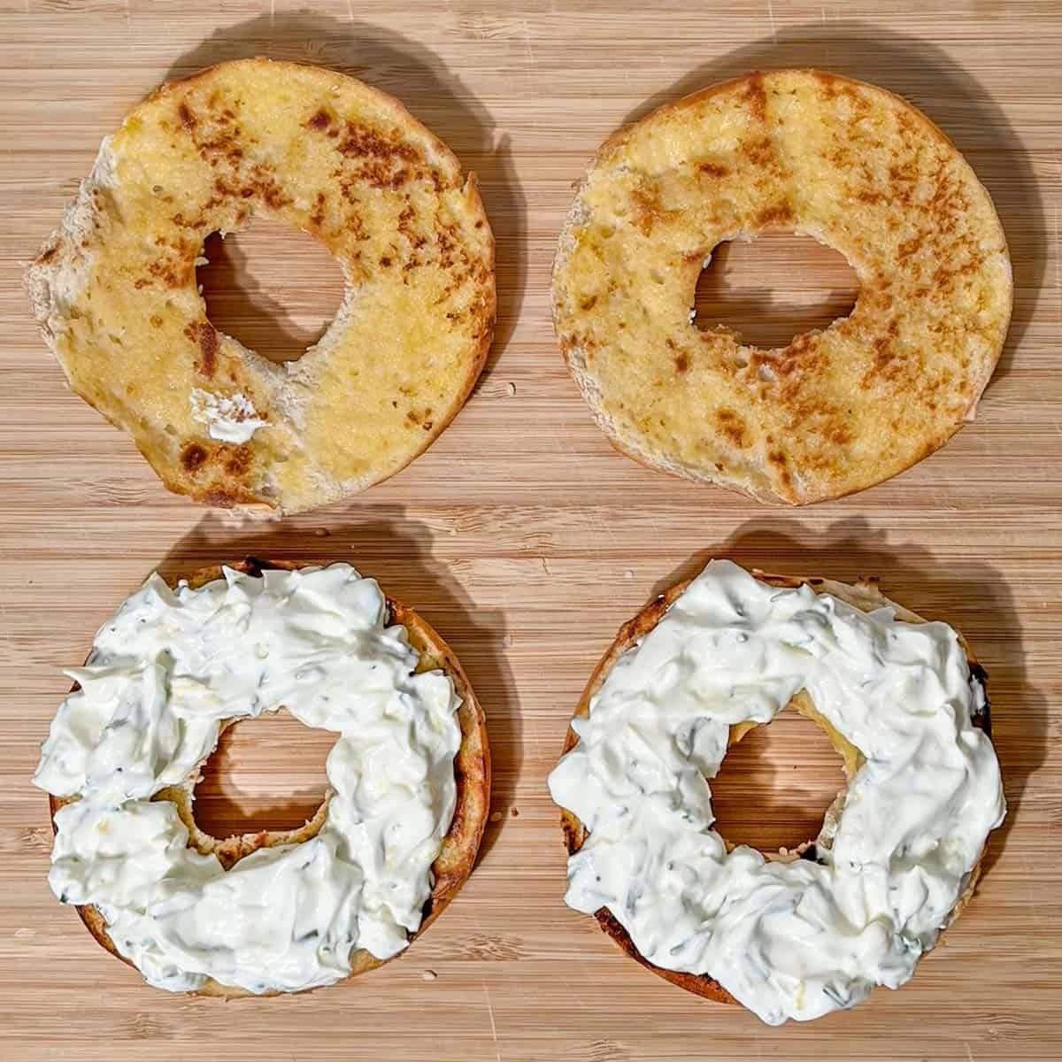 Two bagel halves layered with whipped cream cheese on a wooden board.