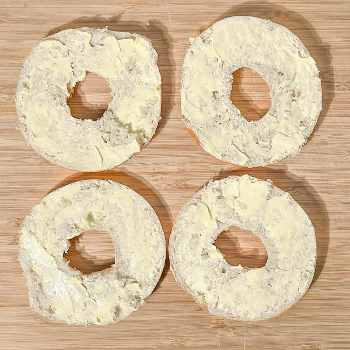 Four bagel halves cut open and spread with butter on a wooden board.