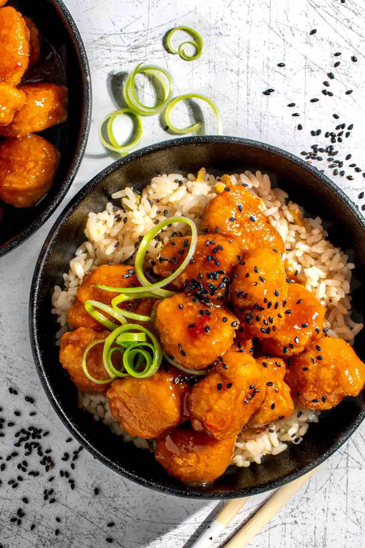 Orange chicken with nuggets on a bed of rice in a black bowl.